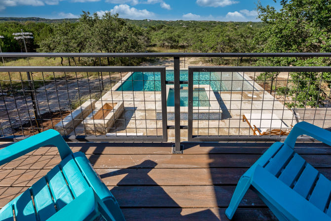 Balcony view with teal deck chairs overlooking a stone patio, rectangular swimming pool and raised spa, wooded hills beyond.