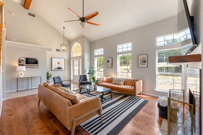 Sunlit modern living room with vaulted ceiling and wooden fan, tan leather sofas, black chairs, striped rug, and tall arched and rectangular windows