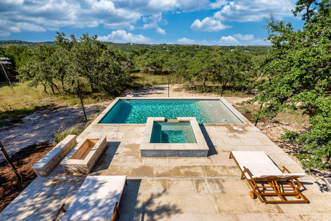 Sunlit aerial view of a modern rectangular outdoor pool with a raised square spa on a travertine patio, wooden lounge chairs, surrounding oak trees and rolling green hills under a bright blue sky.