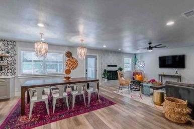 Bright open-concept living and dining room with a long wooden dining table and metal chairs on a fuchsia patterned rug, woven pendant lights, hardwood floors, tiled fireplace, wicker accent chairs, colorful rugs and a wall-mounted TV — modern boho coastal style.