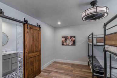 Compact modern farmhouse bedroom with metal bunk beds, rustic sliding barn door opening to a patterned-tile bathroom, hardwood floors, floral wall art and a circular ceiling fan light.