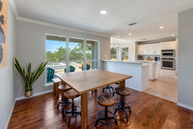 Sunlit open-concept dining area with a large wooden table and industrial swivel stools on hardwood floors, adjacent white kitchen with island, and wide windows framing a backyard pool and trees.