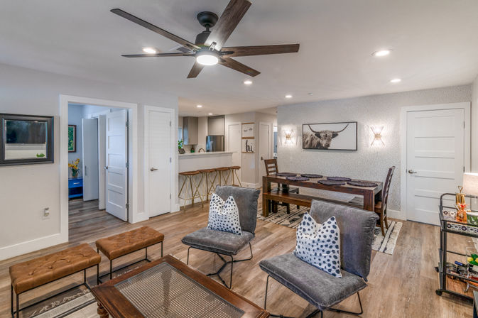 Cozy modern open-concept living and dining area with wood floors, large ceiling fan, two gray lounge chairs with polka-dot pillows, leather ottomans, wooden coffee table, long dining table with bench seating, breakfast bar with three brass stools, and framed highland cow wall art.