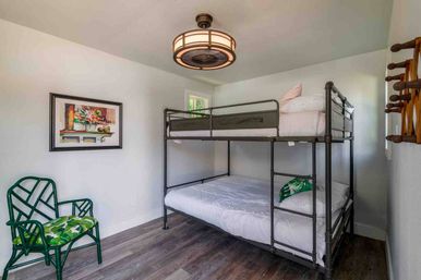 Cozy bright guest room with metal twin-over-full bunk bed, tropical-green accent chair with leafy cushion, wood-look flooring, framed floral art, and round ceiling fan light.