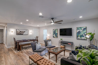 Open-concept modern living and dining room with wood-look floors, gray seating and tufted ottoman, wall-mounted TV, wooden dining table with bench, ceiling fan, botanical artwork and potted plants