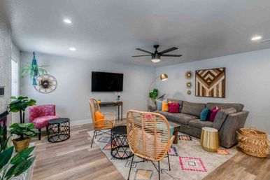 Cozy modern living room interior with a gray sectional sofa, colorful accent pillows, rattan lounge chairs, patterned area rug, wall-mounted TV, black ceiling fan, wood-look floors, indoor plants, and eclectic wall decor.