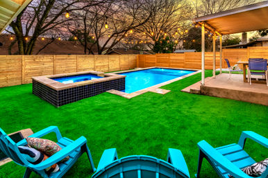Suburban backyard at dusk with rectangular swimming pool and raised tiled hot tub, vibrant green turf, turquoise lounge chairs, covered patio dining area, wood privacy fence and warm string lights overhead.
