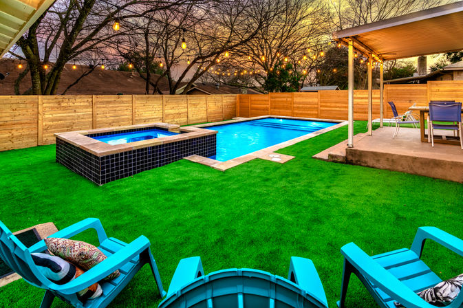 Suburban backyard at dusk with rectangular swimming pool and raised tiled hot tub, vibrant green turf, turquoise lounge chairs, covered patio dining area, wood privacy fence and warm string lights overhead.