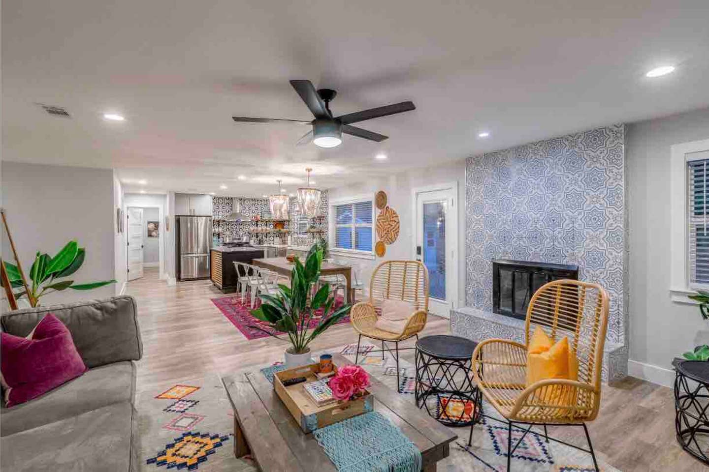 Open-concept living area and kitchen with boho-modern styling — rattan accent chairs with yellow pillow, patterned tile fireplace, wooden coffee table with plants and pink flowers, kitchen island with pendant lights and stainless steel refrigerator, light wood floors and colorful rugs.