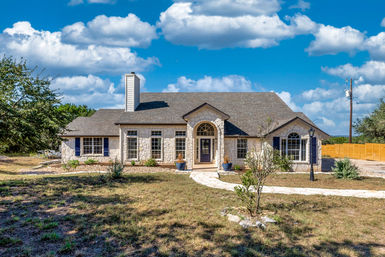 Exterior of a single-story stone ranch-style home with arched front entry, blue shutters, large windows, curved stone walkway and lamppost in a dry front yard under a bright blue sky