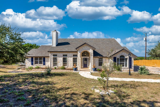 Exterior of a single-story stone ranch-style home with arched front entry, blue shutters, large windows, curved stone walkway and lamppost in a dry front yard under a bright blue sky