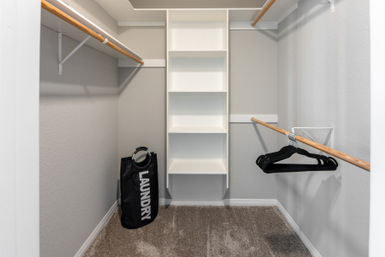 Carpeted walk-in closet with white built-in shelving, wooden hanging rods, a black laundry bag and black hangers in a neutral-toned modern home.
