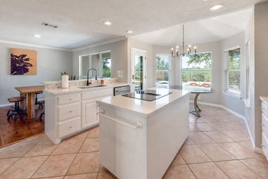 Sunlit open-concept kitchen with a white island and cooktop, bright cabinets, tiled floor, and a bay-window breakfast nook overlooking backyard trees.