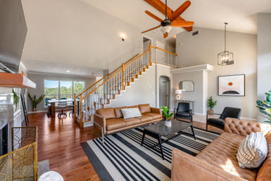 Bright, airy two-story open-plan living room in a suburban home with vaulted ceiling and large windows, wooden staircase, leather sofas and tufted couch, black coffee table on a striped rug, hardwood floors, ceiling fan, and modern minimalist decor.
