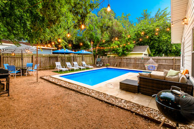 Cozy suburban backyard with rectangular swimming pool at dusk, warm string lights overhead, wicker outdoor sofa with cushions, row of white lounge chairs, umbrella-shaded table, hanging hammock chair and charcoal grill.