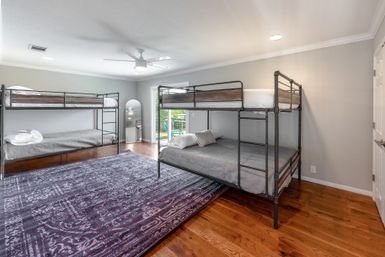 Spacious modern bedroom with two metal bunk beds and gray bedding, warm hardwood floors, large patterned area rug, ceiling fan, and sliding glass door opening to a sunny backyard