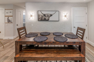 Rustic wooden dining table with six dark round placemats, ladder-back chairs and bench seating in a modern dining room with dotted accent wallpaper, framed highland cow print and twin wall sconces.