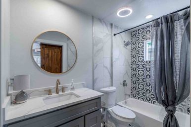 Modern gray bathroom with vanity and round gold mirror, brass faucet, toilet and bathtub featuring black-and-white patterned tile, marble-look walls and knotted gray shower curtain.