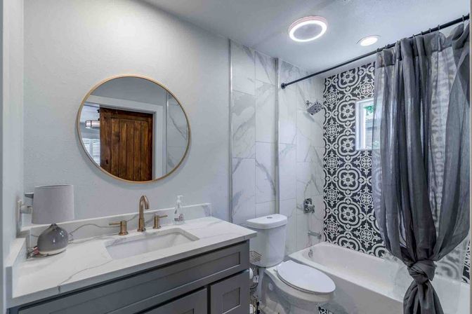 Modern gray bathroom with vanity and round gold mirror, brass faucet, toilet and bathtub featuring black-and-white patterned tile, marble-look walls and knotted gray shower curtain.