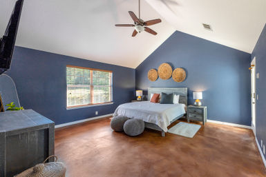 Spacious master bedroom with vaulted ceiling and navy-blue walls, large window with blinds, wooden ceiling fan, large bed with white linens, woven rattan wall decor above the headboard, matching gray nightstands with lamps, knitted poufs and polished brown concrete floor.
