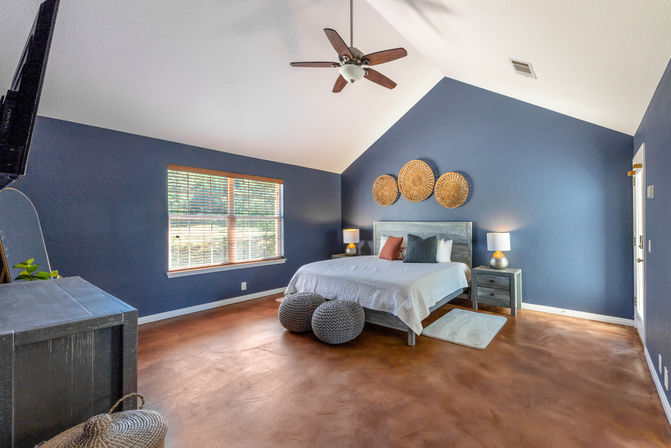 Spacious master bedroom with vaulted ceiling and navy-blue walls, large window with blinds, wooden ceiling fan, large bed with white linens, woven rattan wall decor above the headboard, matching gray nightstands with lamps, knitted poufs and polished brown concrete floor.