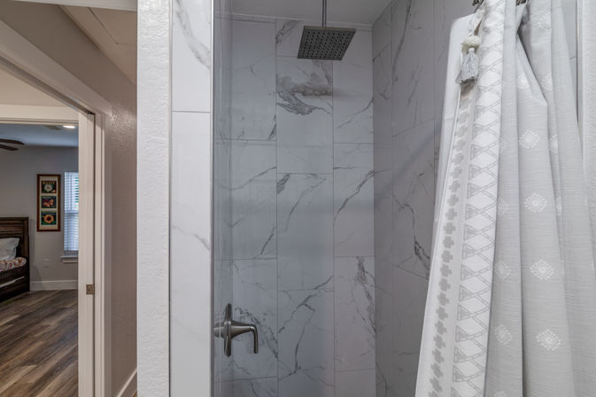 Spa-like home shower with marble-look tile walls, square rain showerhead, brushed-nickel handle and white patterned shower curtain, bedroom visible through open doorway.