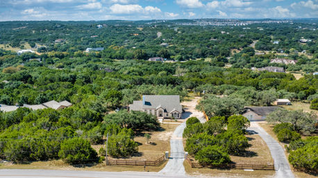 Aerial view of a peaceful country home with a curved private driveway and wooden fenced entrance, surrounded by dense trees and rolling green hills under a blue sky.