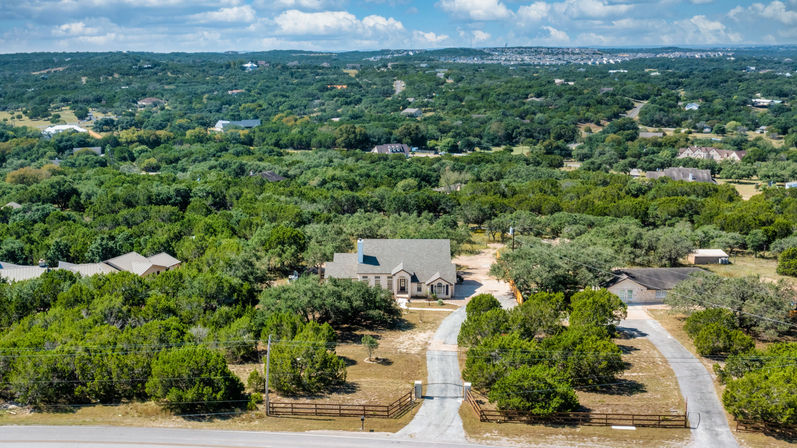Aerial view of a peaceful country home with a curved private driveway and wooden fenced entrance, surrounded by dense trees and rolling green hills under a blue sky.