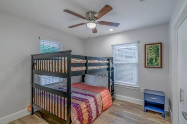 Cozy modern guest bedroom with dark wood bunk bed, colorful quilted bedding, ceiling fan, two windows with blinds, blue nightstand, and wood flooring.