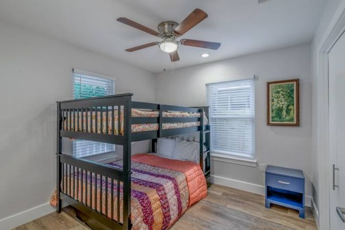 Cozy modern guest bedroom with dark wood bunk bed, colorful quilted bedding, ceiling fan, two windows with blinds, blue nightstand, and wood flooring.
