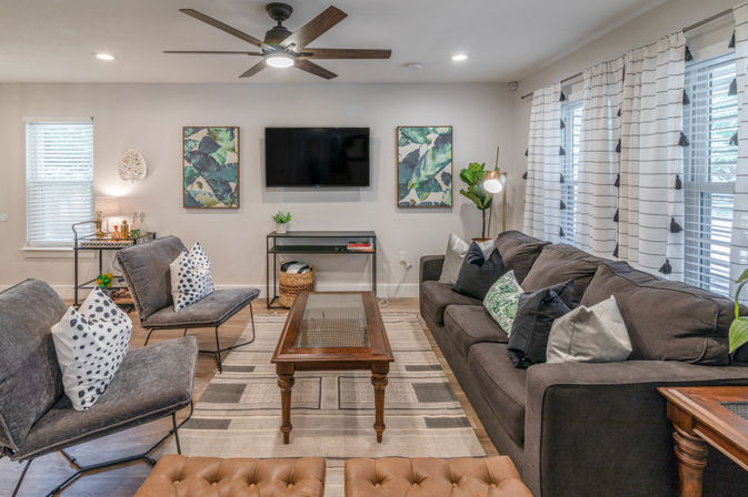 Cozy contemporary living room with dark gray sofa, two gray accent chairs, tufted leather ottoman, wooden coffee table on a patterned rug, wall-mounted TV flanked by botanical prints, ceiling fan, striped curtains, hardwood floor and potted plant.