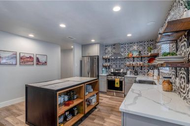 Contemporary kitchen with black-and-white patterned tile backsplash, stainless steel refrigerator and range, large central island with open wooden shelving, marble-look countertops, floating wood shelves and recessed lighting.