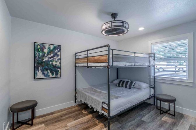 Cozy modern bedroom with industrial metal bunk bed, wood-look plank flooring, abstract green-blue wall art, two round stools, window blinds, and a circular ceiling fan light