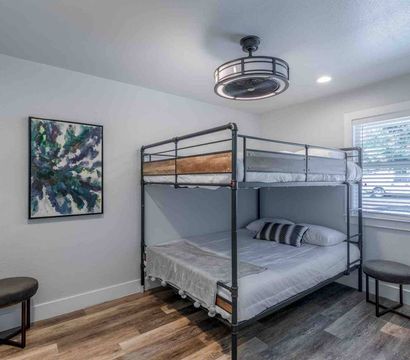 Cozy modern bedroom with industrial metal bunk bed, wood-look plank flooring, abstract green-blue wall art, two round stools, window blinds, and a circular ceiling fan light