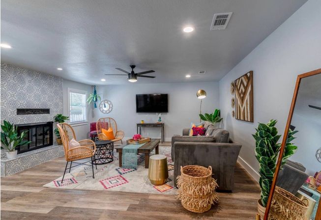 Cozy contemporary living room with patterned-tile fireplace, wall-mounted TV, gray sofa, rattan chairs, colorful pillows and rug, indoor plants, woven baskets and wood-look floors.