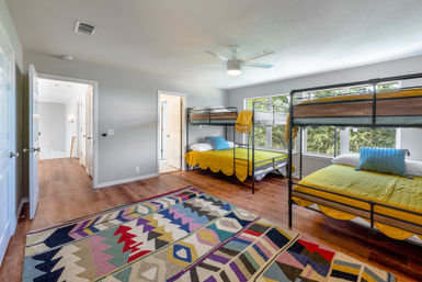 Cheerful sunlit bedroom with two metal bunk bed frames dressed in yellow bedding, large windows overlooking leafy trees, hardwood floors, a colorful geometric area rug, and a modern ceiling fan.