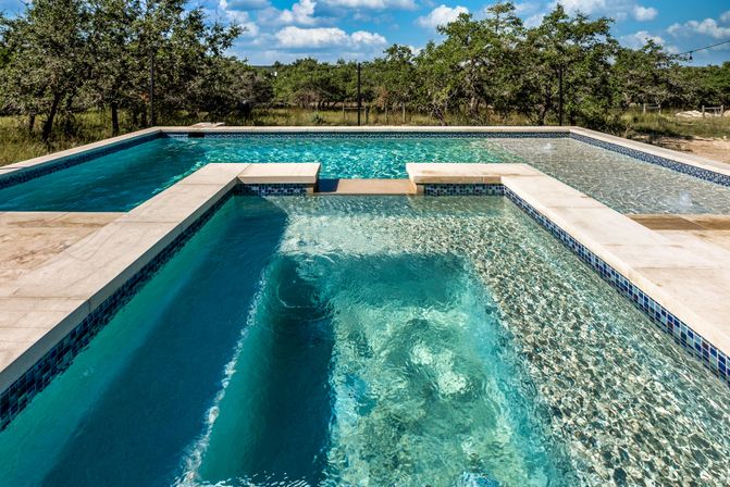 Modern rectangular backyard pool with built-in spa and tanning ledge, crystal turquoise water and tree-lined rural landscape under a blue sky