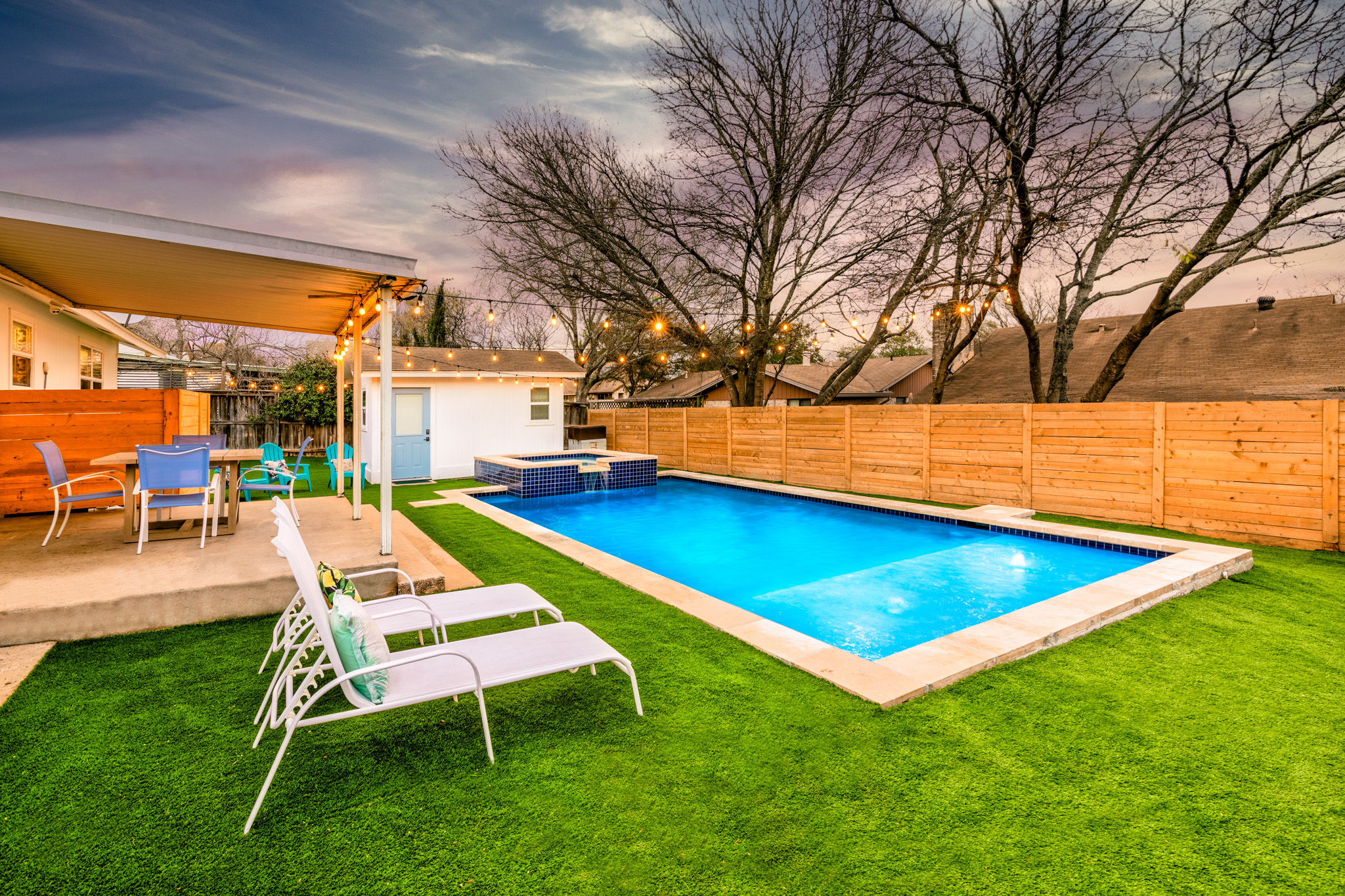 Inviting suburban backyard at dusk with a glowing rectangular pool and raised spa, artificial turf lawn, two white lounge chairs, covered patio with colorful dining chairs, wooden privacy fence and string lights