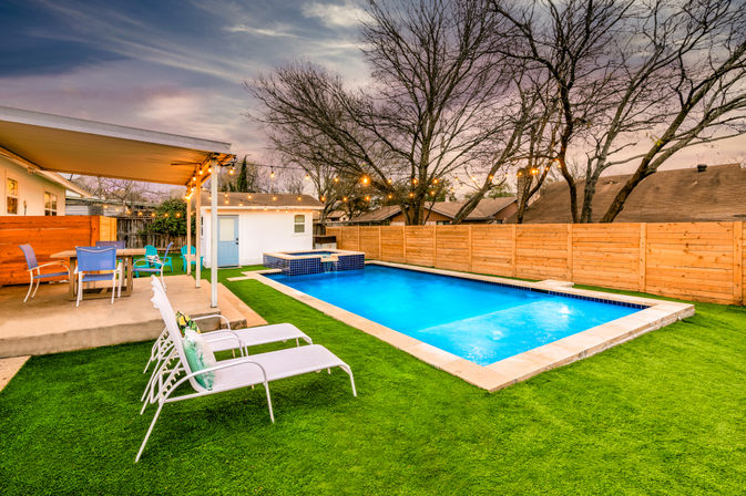 Inviting suburban backyard at dusk with a glowing rectangular pool and raised spa, artificial turf lawn, two white lounge chairs, covered patio with colorful dining chairs, wooden privacy fence and string lights
