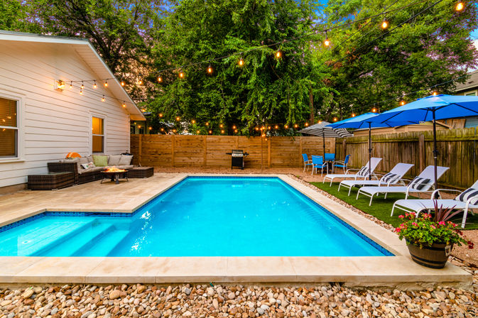 Inviting backyard rectangular swimming pool with turquoise water, twinkle string lights overhead, wicker patio sofa, blue umbrellas, lounge chairs, potted plants and wooden fence