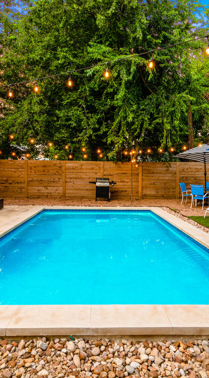 Inviting backyard rectangular swimming pool with turquoise water, twinkle string lights overhead, wicker patio sofa, blue umbrellas, lounge chairs, potted plants and wooden fence