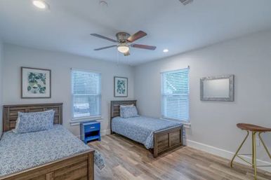 Bright, cozy twin bedroom with two wooden beds in blue patterned bedding, a blue nightstand between two windows with blinds, ceiling fan, botanical wall art and wood-look flooring.