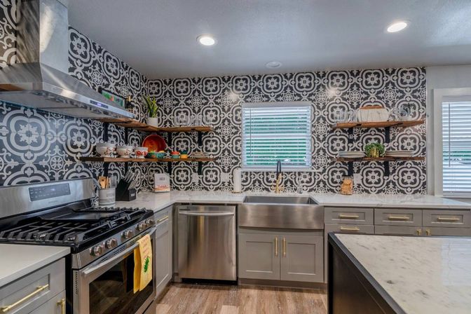 Modern kitchen interior with bold black-and-white patterned tile backsplash, stainless-steel range and dishwasher, farmhouse sink, gray cabinets, open wooden shelves, marble countertops and wood-look floors.