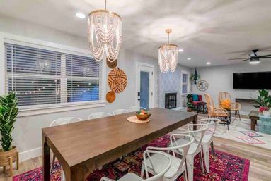 Modern boho open-concept dining and living area with a long wooden table, white metal chairs, beaded chandeliers, colorful Persian-style rug, rattan seating, fireplace, TV, and potted plants.