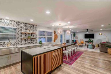 Open-concept modern living space with wood floors: kitchen island featuring slatted wood panels, stainless farmhouse sink and patterned black-and-white tile backsplash, long wooden dining table with metal chairs on a pink rug, pendant chandeliers, recessed lighting and a cozy living area with sofa and wall-mounted TV.