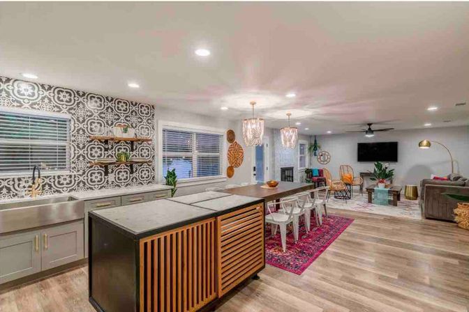 Open-concept modern living space with wood floors: kitchen island featuring slatted wood panels, stainless farmhouse sink and patterned black-and-white tile backsplash, long wooden dining table with metal chairs on a pink rug, pendant chandeliers, recessed lighting and a cozy living area with sofa and wall-mounted TV.