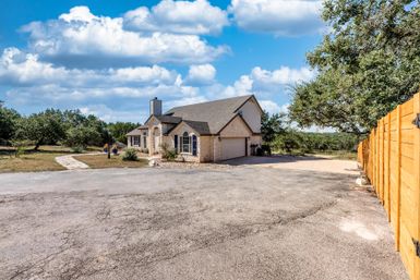 Rural limestone house with attached two-car garage on a wide paved driveway, wooden privacy fence and oak trees, set in open countryside under a bright blue sky with fluffy white clouds.