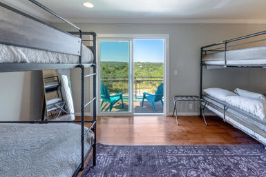 Sunny bunk-room with black metal bunk beds, hardwood floors, sliding glass doors leading to a balcony with turquoise chairs and a sweeping green hillside view.