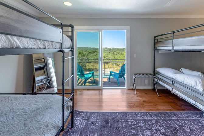 Sunny bunk-room with black metal bunk beds, hardwood floors, sliding glass doors leading to a balcony with turquoise chairs and a sweeping green hillside view.