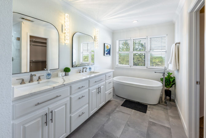 Bright modern white bathroom with double vanity and marble countertop, arched mirrors with sparkling wall sconces, freestanding tub beneath sunlit window blinds, potted plant and gray tile floor